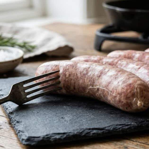 Dettaglio ravvicinato di salsicce fresche intere su un tagliere di ardesia, con una forchetta appoggiata vicino ma che non le punge. Un esempio pratico di come cuocere la salsiccia senza bucarla per mantenerne la succosità.