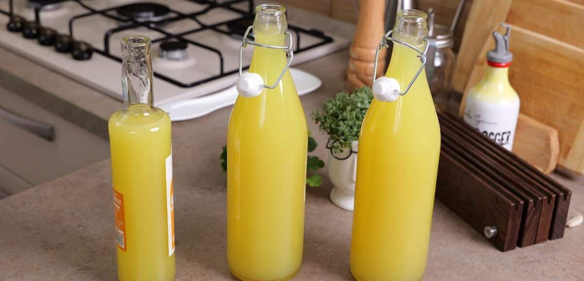 Tre bottiglie di vetro piene di limoncello fatto in casa, posizionate su un piano cucina accanto a utensili e un fornello a gas.