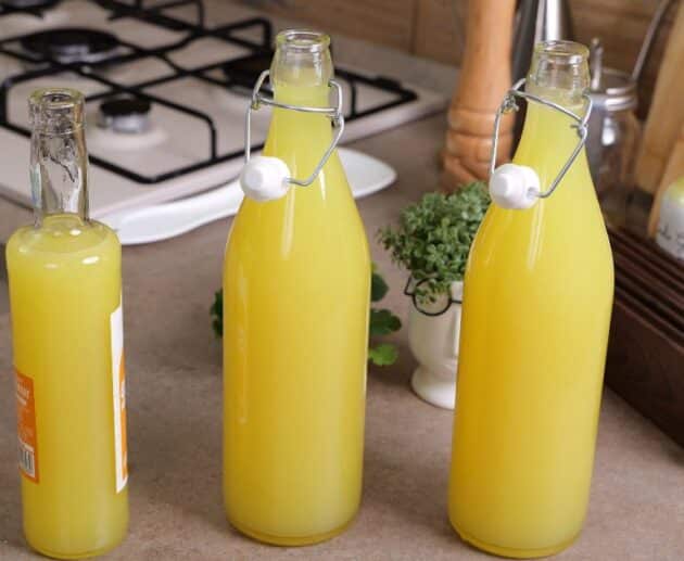 Tre bottiglie di vetro piene di limoncello fatto in casa, posizionate su un piano cucina accanto a utensili e un fornello a gas.