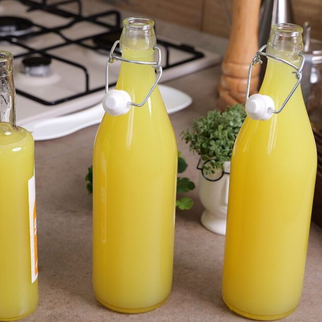 Tre bottiglie di vetro piene di limoncello fatto in casa, posizionate su un piano cucina accanto a utensili e un fornello a gas.