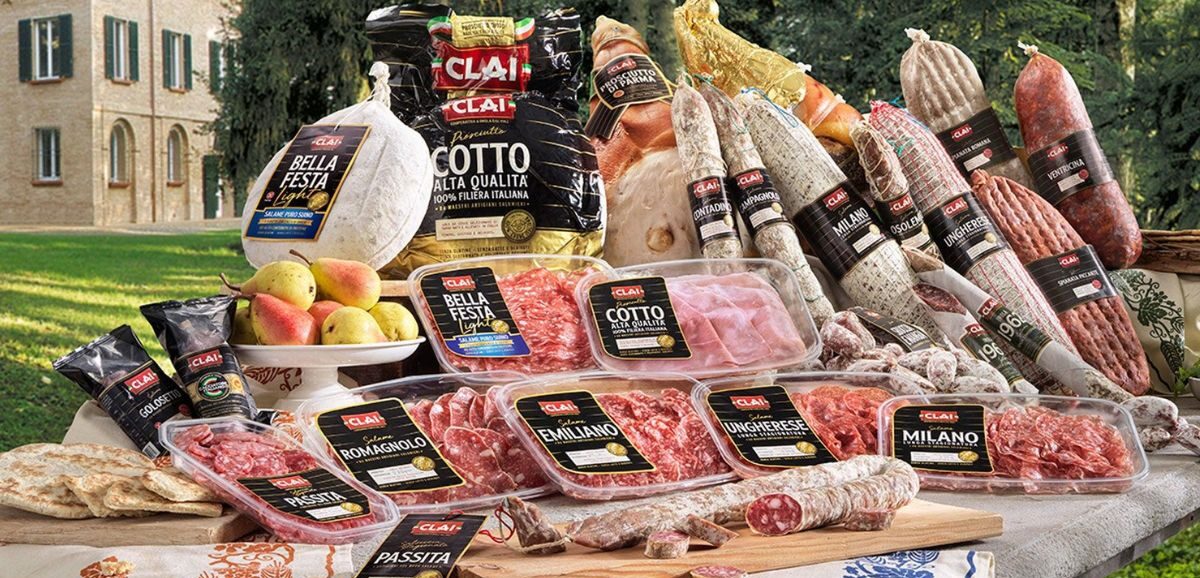 Ricca selezione di salumi e affettati confezionati a marchio CLAI disposti su un tavolo all'aperto davanti a una tenuta storica.